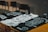 Three black and white graphic t-shirts are laid out on a wooden surface, featuring intricate designs and texts. A dark-colored square bottle, possibly an alcoholic beverage, is placed amongst the shirts. In the background, there are some unoccupied chairs with a dimly lit atmosphere surrounding the scene.
