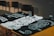 Three black and white graphic t-shirts are laid out on a wooden surface, featuring intricate designs and texts. A dark-colored square bottle, possibly an alcoholic beverage, is placed amongst the shirts. In the background, there are some unoccupied chairs with a dimly lit atmosphere surrounding the scene.