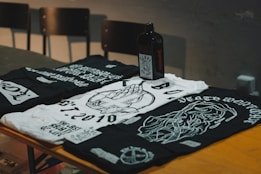 Three black and white graphic t-shirts are laid out on a wooden surface, featuring intricate designs and texts. A dark-colored square bottle, possibly an alcoholic beverage, is placed amongst the shirts. In the background, there are some unoccupied chairs with a dimly lit atmosphere surrounding the scene.