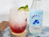 A glass filled with a refreshing iced beverage, containing crushed ice, a red liquid and garnished with a slice of lime and mint leaves. Beside the glass is a bottle of Mineragua sparkling water, which is clear with a blue label featuring bubbles.