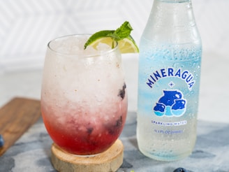 A glass filled with a refreshing iced beverage, containing crushed ice, a red liquid and garnished with a slice of lime and mint leaves. Beside the glass is a bottle of Mineragua sparkling water, which is clear with a blue label featuring bubbles.
