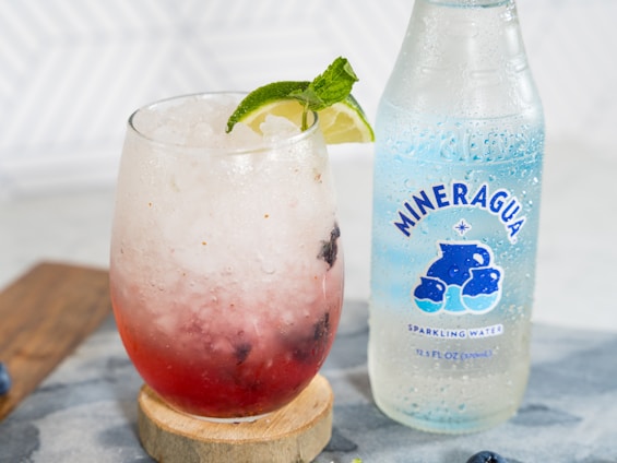 A glass filled with a refreshing iced beverage, containing crushed ice, a red liquid and garnished with a slice of lime and mint leaves. Beside the glass is a bottle of Mineragua sparkling water, which is clear with a blue label featuring bubbles.