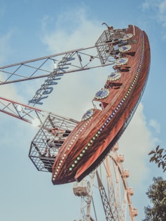 A large amusement park ride shaped like a pirate ship swings through the air, set against a background of blue sky and scattered clouds. The ship is adorned with colorful lights and decorative elements. People are visible inside the ride cabin, experiencing the thrill. In the distance, part of a Ferris wheel can be seen.
