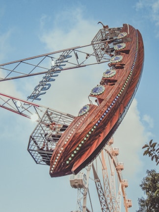 A large amusement park ride shaped like a pirate ship swings through the air, set against a background of blue sky and scattered clouds. The ship is adorned with colorful lights and decorative elements. People are visible inside the ride cabin, experiencing the thrill. In the distance, part of a Ferris wheel can be seen.