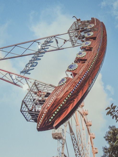 A large amusement park ride shaped like a pirate ship swings through the air, set against a background of blue sky and scattered clouds. The ship is adorned with colorful lights and decorative elements. People are visible inside the ride cabin, experiencing the thrill. In the distance, part of a Ferris wheel can be seen.