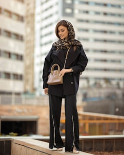 A woman stands confidently against an urban backdrop, wearing a black coat and pants with a patterned headscarf. She holds a small brown handbag and appears poised and stylish.