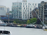 A riverside scene features modern apartment buildings and older brick structures. Several moored boats line the waterfront, including a mix of sailboats and houseboats. The surrounding area has some industrial elements and greenery along the shore.