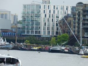 A riverside scene features modern apartment buildings and older brick structures. Several moored boats line the waterfront, including a mix of sailboats and houseboats. The surrounding area has some industrial elements and greenery along the shore.