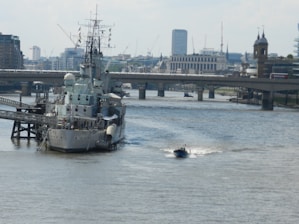 A large naval ship is moored on a river alongside a cityscape backdrop with modern and historical buildings. A small motorboat speeds past in the foreground, creating a white wake behind it. A bridge spans the river in the background, with traffic moving across it.