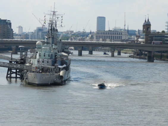 A large naval ship is moored on a river alongside a cityscape backdrop with modern and historical buildings. A small motorboat speeds past in the foreground, creating a white wake behind it. A bridge spans the river in the background, with traffic moving across it.