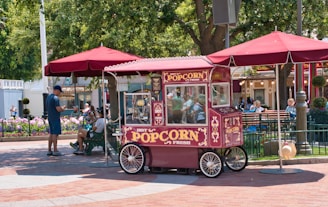 A vintage-style popcorn cart with ornate decorations sits under a maroon umbrella. The cart is surrounded by people, including a man in blue and others seated on a bench in a park-like setting. Colorful flowers and trees enhance the lively atmosphere.