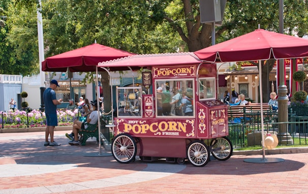 A vintage-style popcorn cart with ornate decorations sits under a maroon umbrella. The cart is surrounded by people, including a man in blue and others seated on a bench in a park-like setting. Colorful flowers and trees enhance the lively atmosphere.