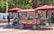 A vintage-style popcorn cart with ornate decorations sits under a maroon umbrella. The cart is surrounded by people, including a man in blue and others seated on a bench in a park-like setting. Colorful flowers and trees enhance the lively atmosphere.