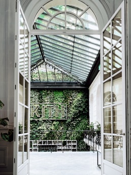 An elegant indoor space with a large glass ceiling and white framed glass doors leading to a lush vertical garden wall with the word 'KITH' displayed in a sign. The space exudes a modern aesthetic, combining natural greenery with architectural elements.