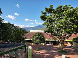 A scenic view of a landscaped area with lush green trees and a building with a red-tiled roof against a backdrop of mountains and a clear blue sky. A few people are visible walking near the building, and a sign with the text 'Rio lento' is present.