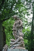 A bronze statue of a young woman standing gracefully, with one hand holding a bouquet of flowers and the other hand at her side. In front of her, a figure of a man appears to be in a protective or comforting pose. The statue is placed outdoors, surrounded by lush green trees and foliage. At the base of the statue, there are flowers and a wreath with ribbons.