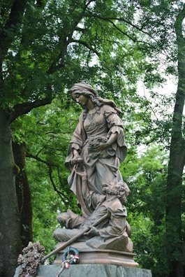 A bronze statue of a young woman standing gracefully, with one hand holding a bouquet of flowers and the other hand at her side. In front of her, a figure of a man appears to be in a protective or comforting pose. The statue is placed outdoors, surrounded by lush green trees and foliage. At the base of the statue, there are flowers and a wreath with ribbons.