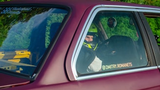 A person wearing a helmet is seated inside a car, visible through the rear side window. The car is maroon-colored and has a sticker on the window with a social media handle. There is greenery reflected on the window and a bright yellow and black roll cage is visible inside the car.