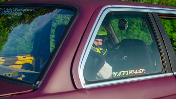 A person wearing a helmet is seated inside a car, visible through the rear side window. The car is maroon-colored and has a sticker on the window with a social media handle. There is greenery reflected on the window and a bright yellow and black roll cage is visible inside the car.