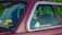A person wearing a helmet is seated inside a car, visible through the rear side window. The car is maroon-colored and has a sticker on the window with a social media handle. There is greenery reflected on the window and a bright yellow and black roll cage is visible inside the car.