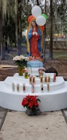 A religious statue of the Virgin Mary stands on a white pedestal, surrounded by lit devotional candles and flowers, including red poinsettias and white roses. Balloons in red, white, and green hover above, and the background features trees with hanging moss.