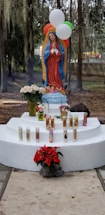 A religious statue of the Virgin Mary stands on a white pedestal, surrounded by lit devotional candles and flowers, including red poinsettias and white roses. Balloons in red, white, and green hover above, and the background features trees with hanging moss.