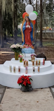 A religious statue of the Virgin Mary stands on a white pedestal, surrounded by lit devotional candles and flowers, including red poinsettias and white roses. Balloons in red, white, and green hover above, and the background features trees with hanging moss.