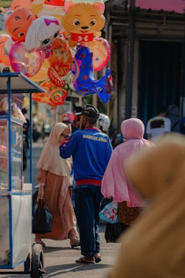 People are gathered around a street vendor selling colorful balloons shaped like animals and cartoon characters. A man wearing a blue jacket and a cap is interacting with two women dressed in modest clothing. The setting appears to be a market or street fair, with other people and stalls visible in the background.