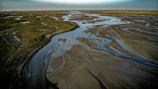 An expansive landscape with a winding network of shallow water channels flowing through a flat, sandy terrain. Green vegetation is visible sparsely distributed on one side, while the other side is largely barren. The distant horizon merges into a hazy, blue sky, suggesting an open and vast environment.