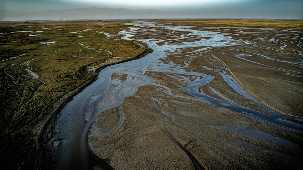 An expansive landscape with a winding network of shallow water channels flowing through a flat, sandy terrain. Green vegetation is visible sparsely distributed on one side, while the other side is largely barren. The distant horizon merges into a hazy, blue sky, suggesting an open and vast environment.