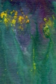 An abstract watercolor painting features a blend of deep green, purple, and scattered bursts of bright yellow. The textured background creates a fluid and organic appearance, with colors merging seamlessly.