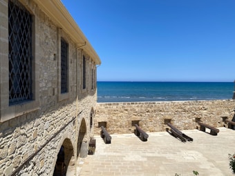 A historic stone building with arched windows stands beside an open courtyard featuring several old cannons pointed towards a calm blue sea under a cloudless sky.