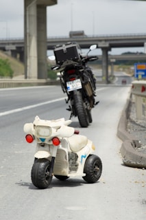 A small toy motorcycle is placed on the roadside of a highway. Behind the toy motorcycle, there is a larger real motorcycle parked on the side of the road. The road is deserted with no other vehicles in sight, and there are overpasses in the background.