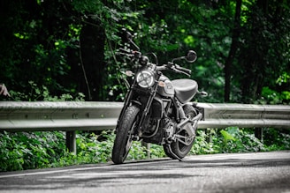 A motorcycle is parked on a winding road surrounded by lush green foliage. The sunlight filters through the trees, casting dappled shadows on the asphalt. The road is bordered by a metal guardrail, and the scene conveys a sense of tranquility and adventure.