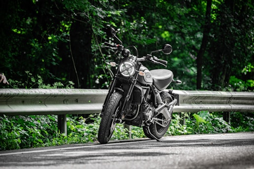 A motorcycle is parked on a winding road surrounded by lush green foliage. The sunlight filters through the trees, casting dappled shadows on the asphalt. The road is bordered by a metal guardrail, and the scene conveys a sense of tranquility and adventure.
