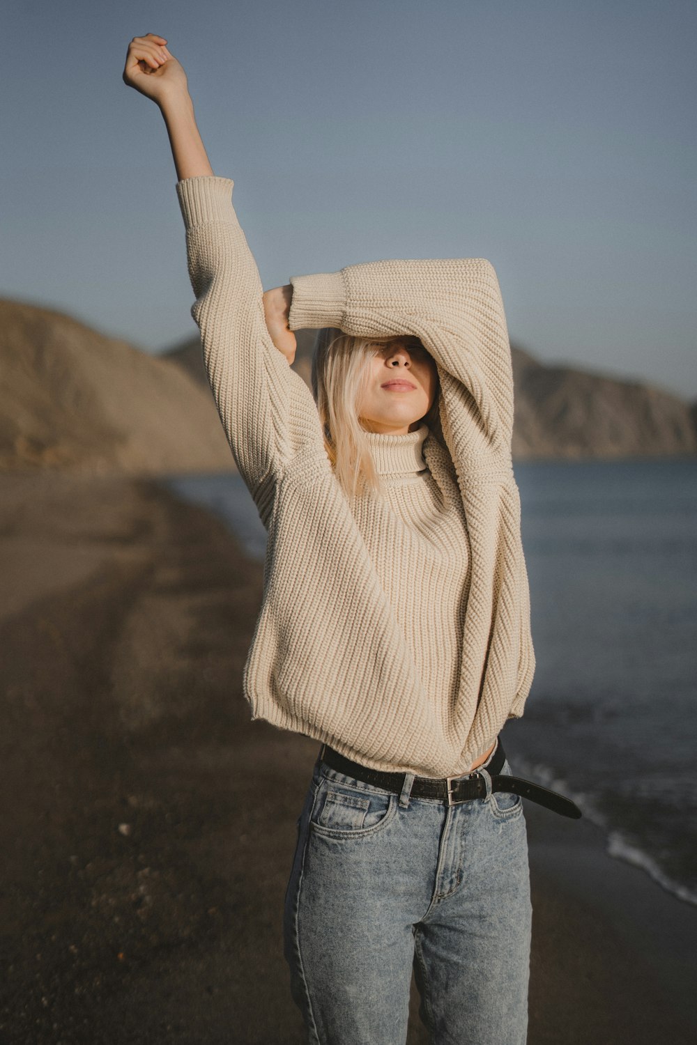 woman in beige knit sweater and blue denim jeans standing on beach shore during daytime
