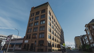 A multi-story brick building with numerous windows is situated on a city street. The building has an old architectural style and features signs indicating spaces for rent. Surrounding it are modern buildings and a bright blue sky.