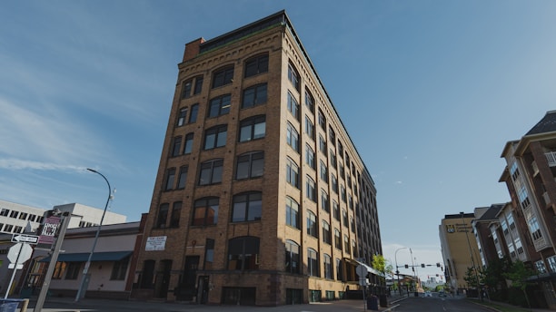 A multi-story brick building with numerous windows is situated on a city street. The building has an old architectural style and features signs indicating spaces for rent. Surrounding it are modern buildings and a bright blue sky.