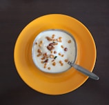 A bowl of yogurt garnished with small fried lentils and nuts sits in the center of a bright yellow plate. A metal spoon is placed beside the bowl, which is positioned on a dark wooden table.