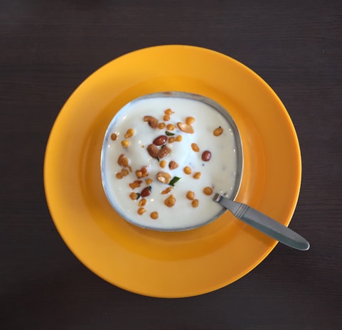 A bowl of yogurt garnished with small fried lentils and nuts sits in the center of a bright yellow plate. A metal spoon is placed beside the bowl, which is positioned on a dark wooden table.
