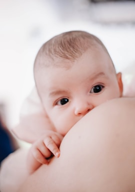 A baby is breastfeeding, with eyes wide open and a gentle expression. The image captures a close-up view with the baby's hand resting on the mother's skin. Soft background enhances the intimate and nurturing atmosphere.