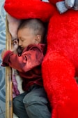 A child sleeps peacefully in what appears to be a small bed or cot, cuddling a large, vivid red teddy bear. The setting is cozy with soft padding, and the child is wearing a deep red shirt. The flooring is visible with a patterned design.