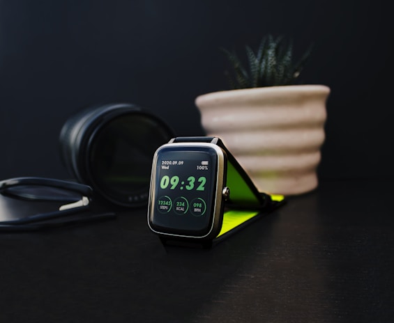 A sleek, modern smartwatch displays the time as 09:32. The screen indicates step count, calories burned, and heart rate. Nearby, a black camera lens and a plant in a beige ribbed pot contribute to the aesthetic setting on a dark surface. There is also a pair of glasses with a black frame.