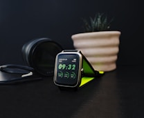 A sleek, modern smartwatch displays the time as 09:32. The screen indicates step count, calories burned, and heart rate. Nearby, a black camera lens and a plant in a beige ribbed pot contribute to the aesthetic setting on a dark surface. There is also a pair of glasses with a black frame.
