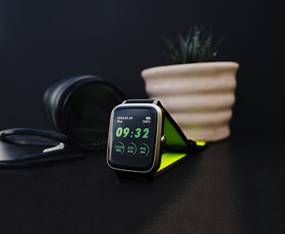 A sleek, modern smartwatch displays the time as 09:32. The screen indicates step count, calories burned, and heart rate. Nearby, a black camera lens and a plant in a beige ribbed pot contribute to the aesthetic setting on a dark surface. There is also a pair of glasses with a black frame.