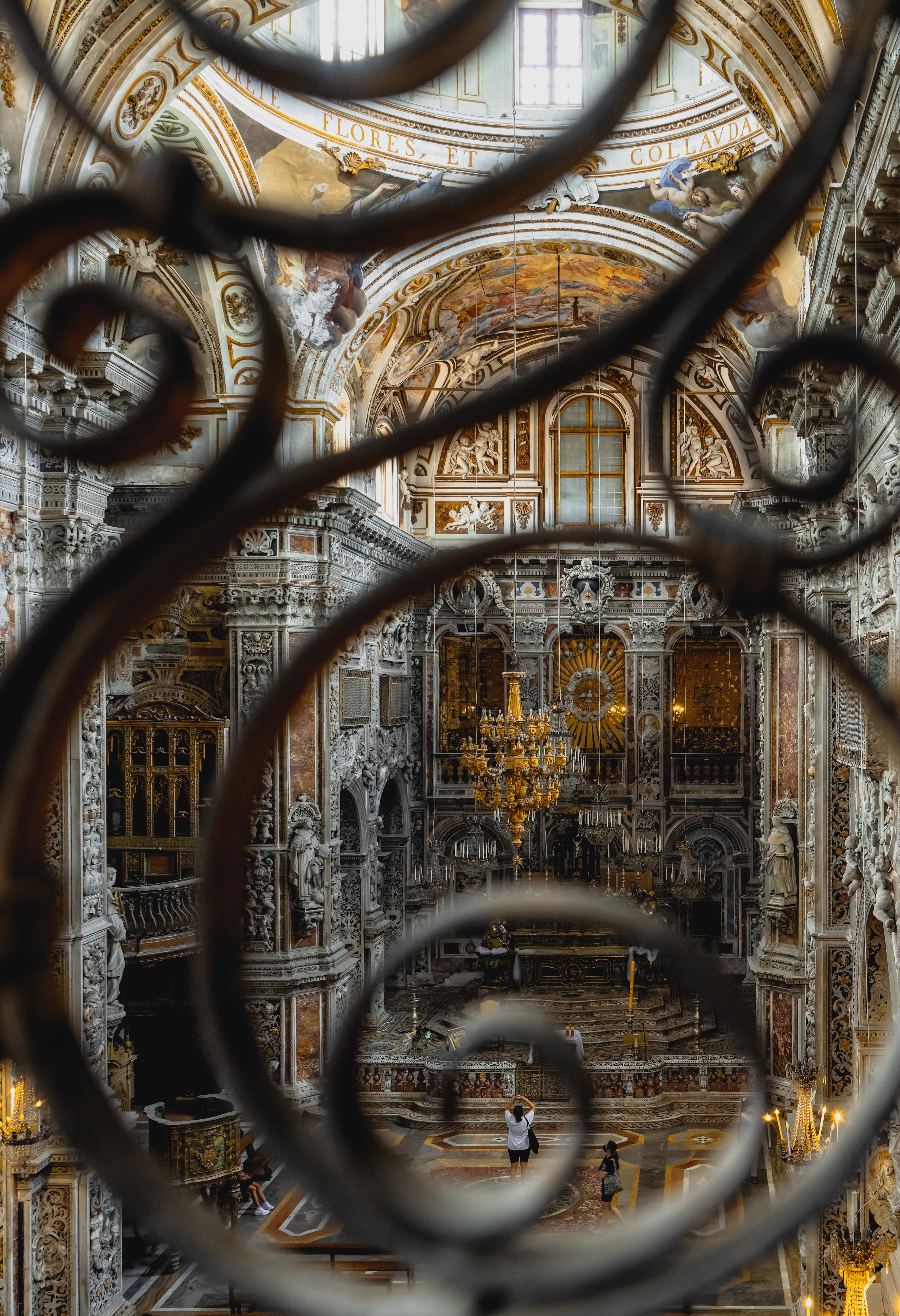 Looking through an ornate wrought-iron screen into a gilded, frescoed baroque church interior with a lit chandelier.