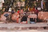 A street scene depicts two individuals engaged in ironing clothes on a makeshift ironing table. A bicycle leans against a wall in the background, and the surrounding area includes trees and hanging cloths. The setting is outdoors with a mix of natural and urban elements.