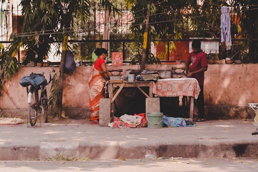 A street scene depicts two individuals engaged in ironing clothes on a makeshift ironing table. A bicycle leans against a wall in the background, and the surrounding area includes trees and hanging cloths. The setting is outdoors with a mix of natural and urban elements.