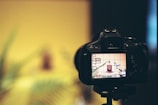 A DSLR camera is positioned on a tripod, capturing an artistic composition that features a small bottle and some plant leaves. The camera's screen displays the scene with a grid overlay, while the background is blurred.