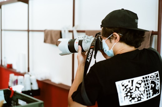 A person wearing a mask and black cap is using a Sony camera with a large lens. The individual is indoors, standing near shelves with various objects, possibly in a professional or creative environment.